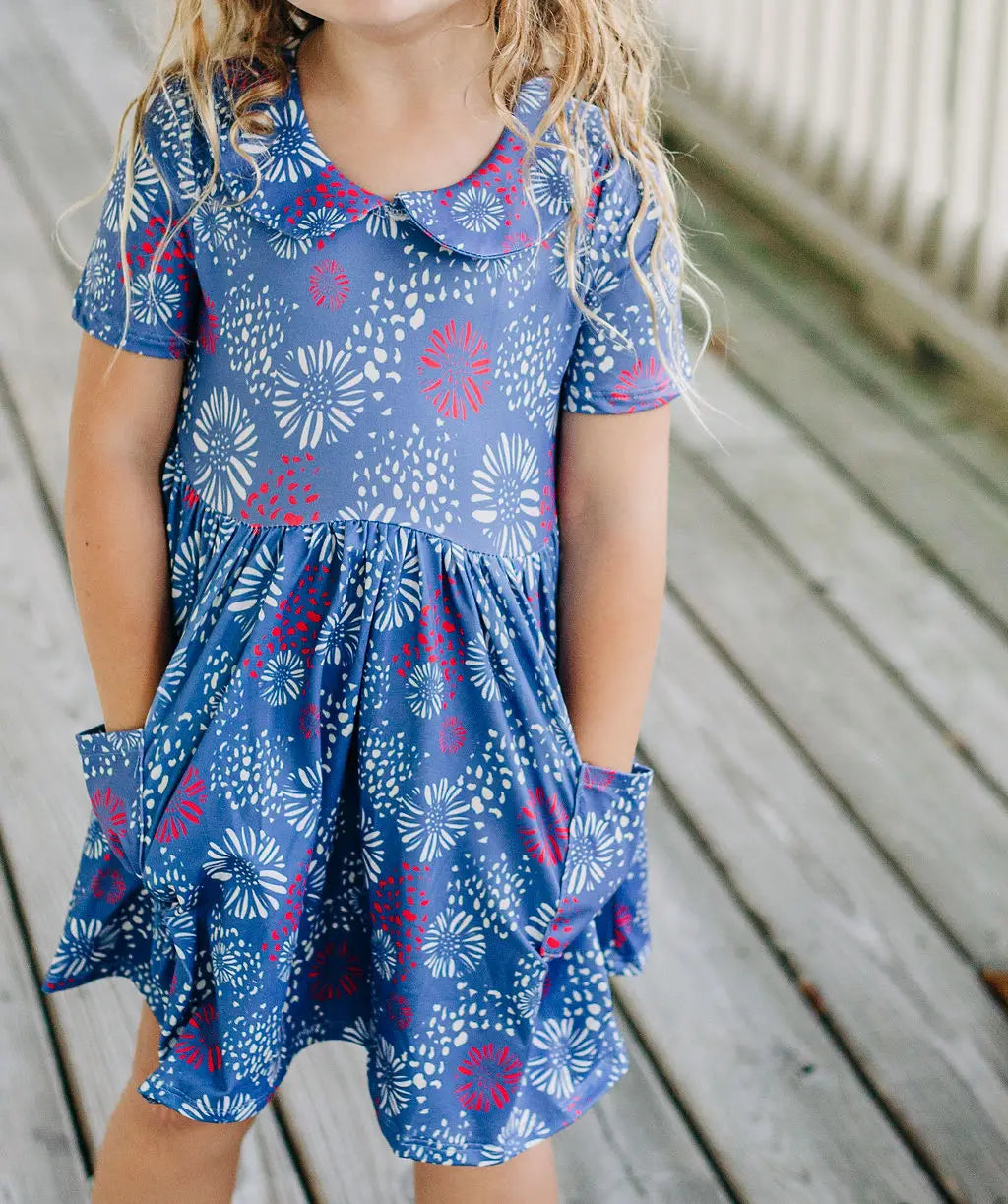 A little girl, wearing the Fireworks Twirl Dress by Sugar Bee Clothing, is standing on a wooden deck.