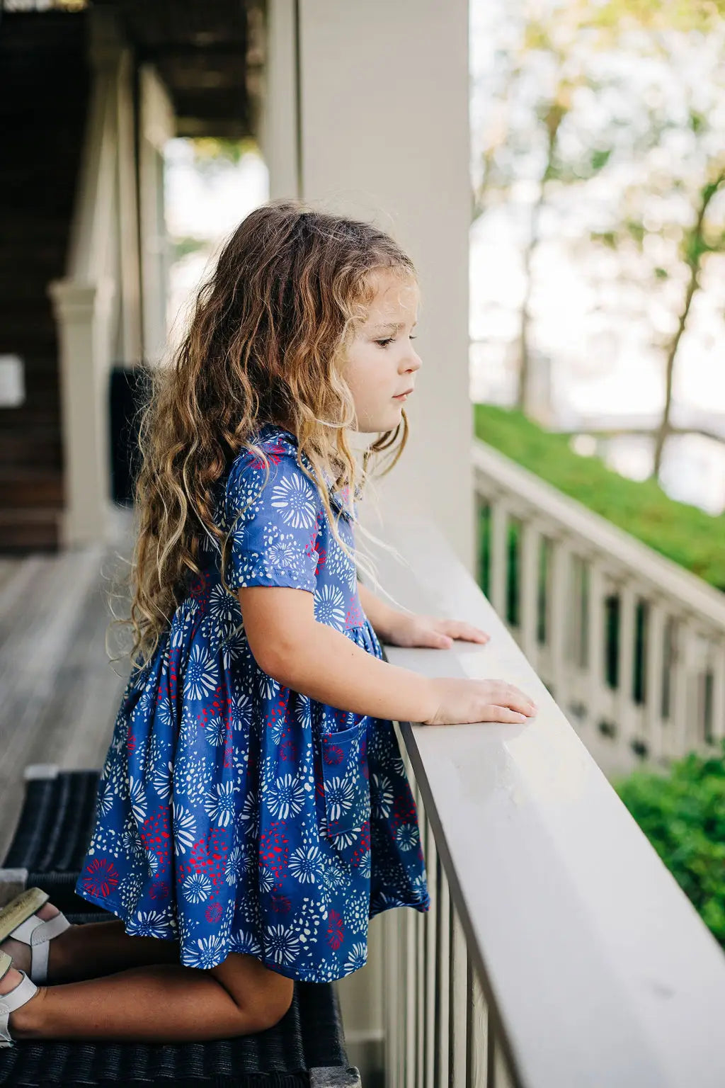 A little girl standing on a porch next to a railing in her Fireworks Twirl Dress by Sugar Bee Clothing.