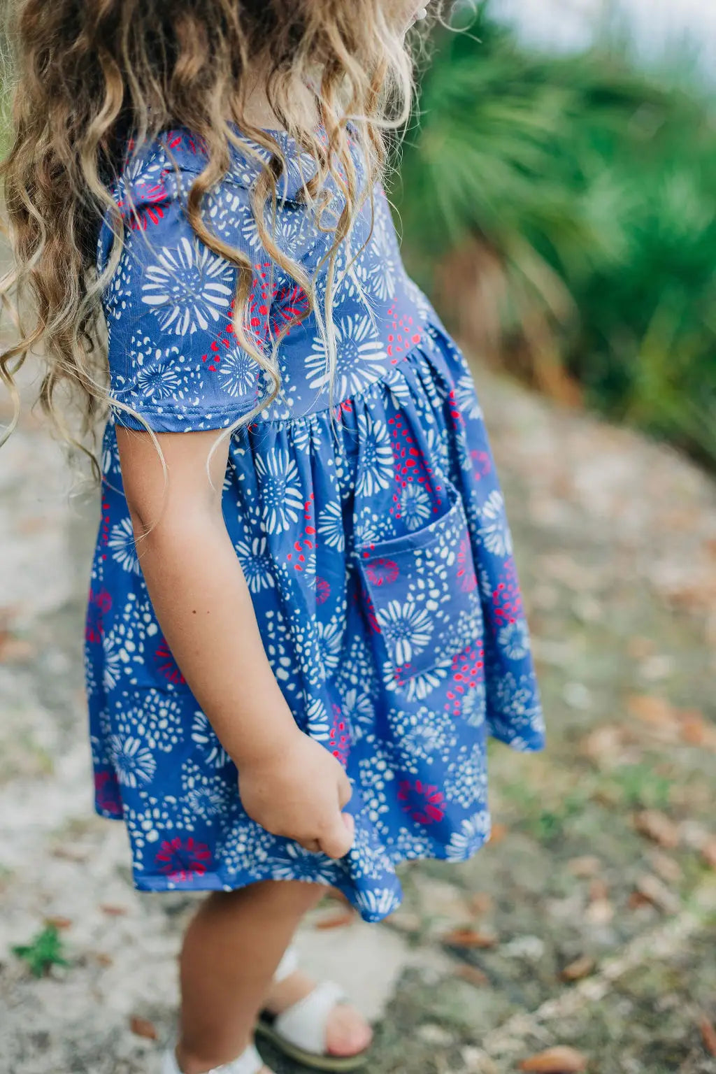 A little girl, wearing the Fireworks Twirl Dress from Sugar Bee Clothing, is standing on a dirt road.