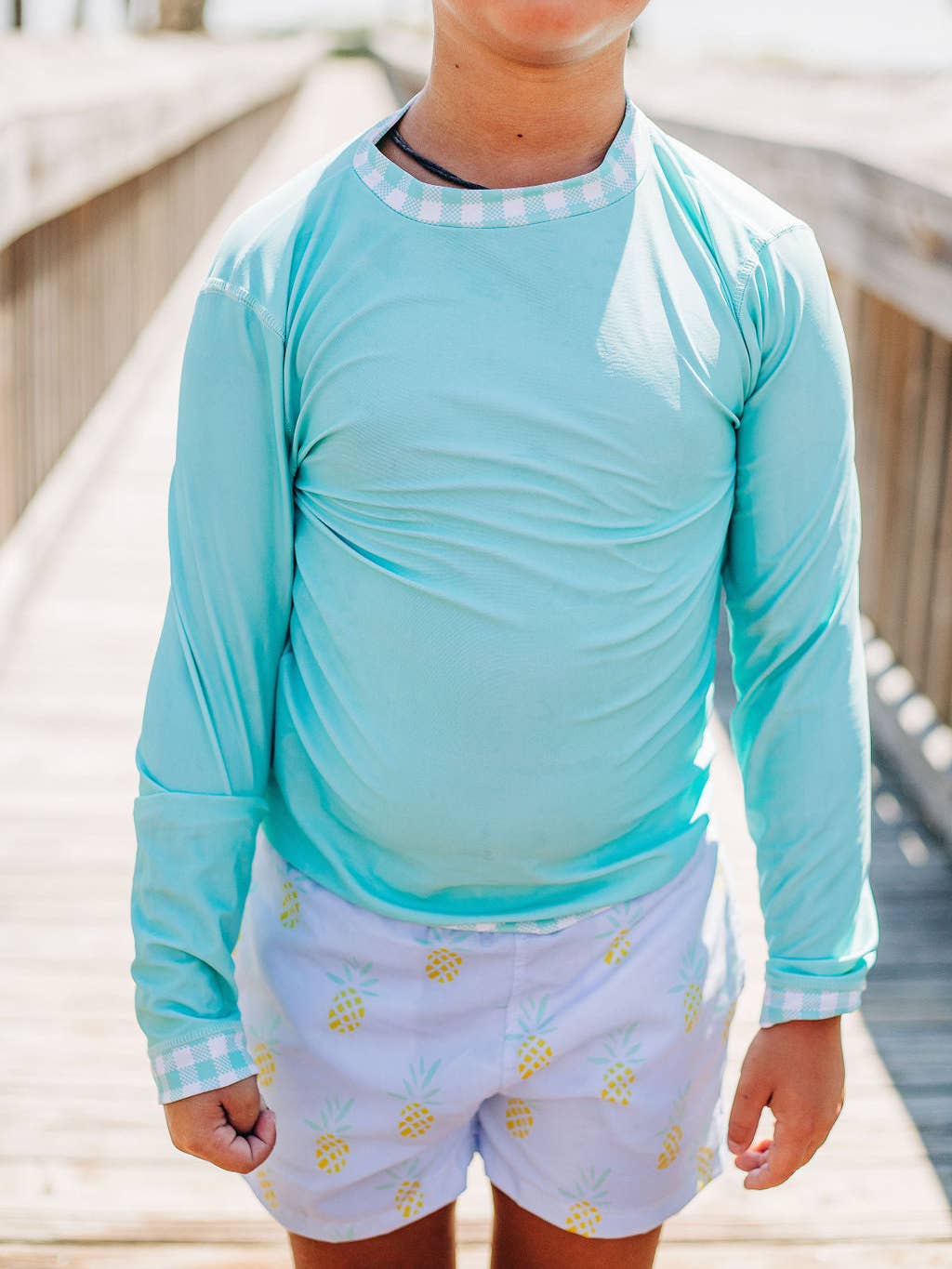 A young boy wearing a Boys Gingham Rashguard by Sugar Bee Clothing in a blue shirt and white shorts.