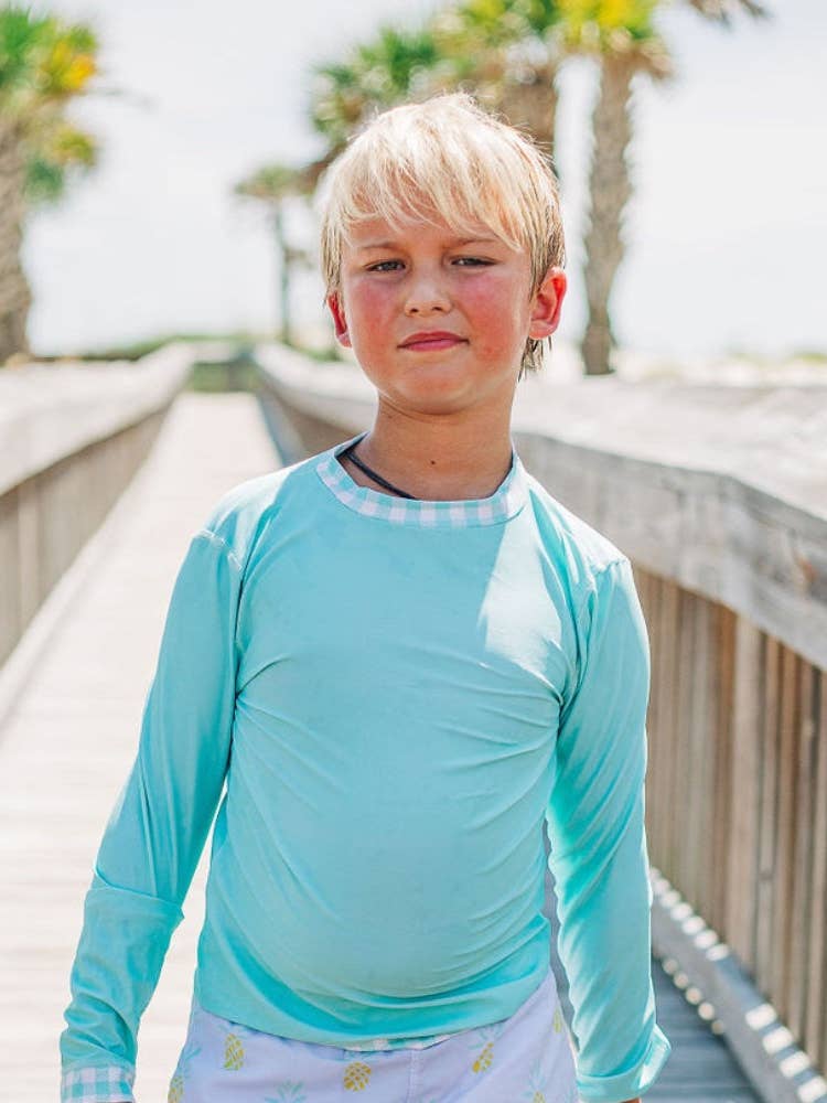 A young boy is standing on a boardwalk wearing a Sugar Bee Clothing Boys Gingham Rashguard.