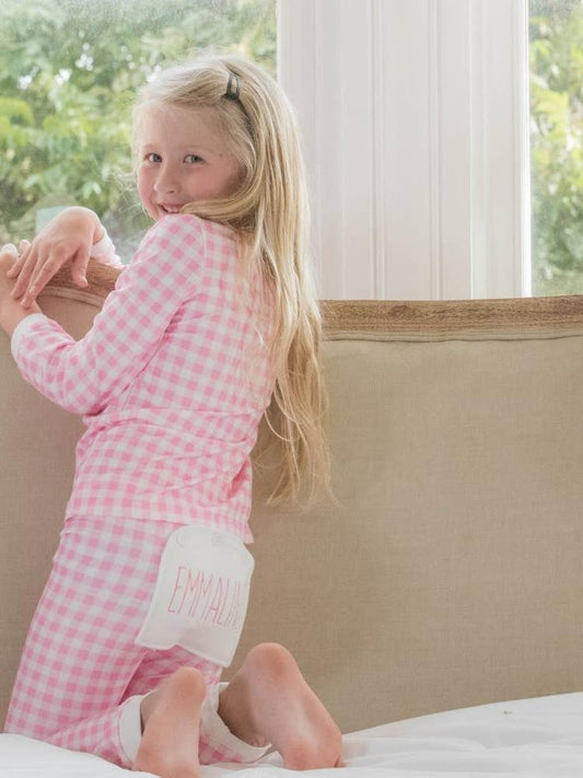 A little girl sitting on a couch wearing Sugar Bee Clothing's Pink Gingham Buttflap Pajamas.