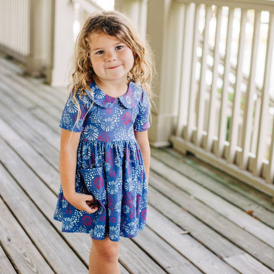 A little girl is standing on a porch wearing the Fireworks Twirl Dress from Sugar Bee Clothing.
