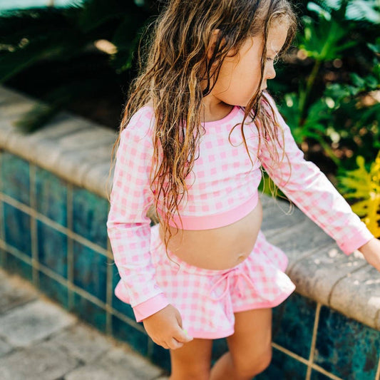 a little girl wearing the Sugar Bee Clothing Pink Gingham Girls Skirt Bikini, standing on a ledge.