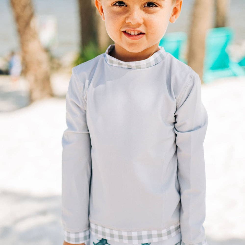 A young boy standing on a beach wearing a Sugar Bee Clothing Boys Gingham - Rashguard wet suit.
