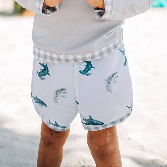 A little boy wearing Sharks - Swim Shorts by Sugar Bee Clothing on the beach.