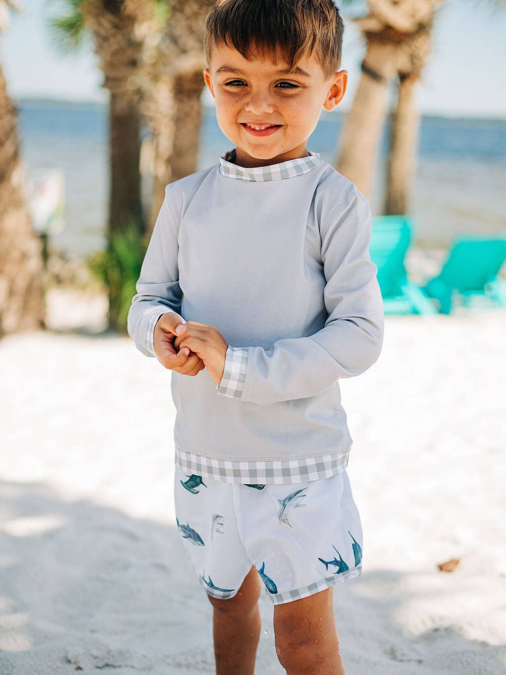 A young boy standing on a beach next to a palm tree, wearing a Boys Gingham Rashguard from Sugar Bee Clothing.