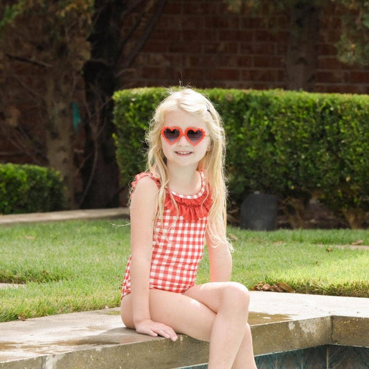 A little girl wearing Sugar Bee Clothing's Red Gingham Bow Girls Swimsuit and sunglasses sitting on a ledge.