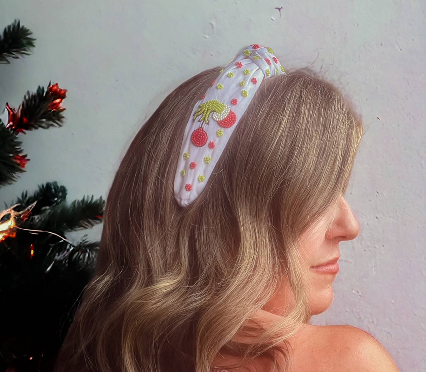 A Festive Beaded Christmas Headband by Bash adds a festive touch to a woman posing in front of a Christmas tree.