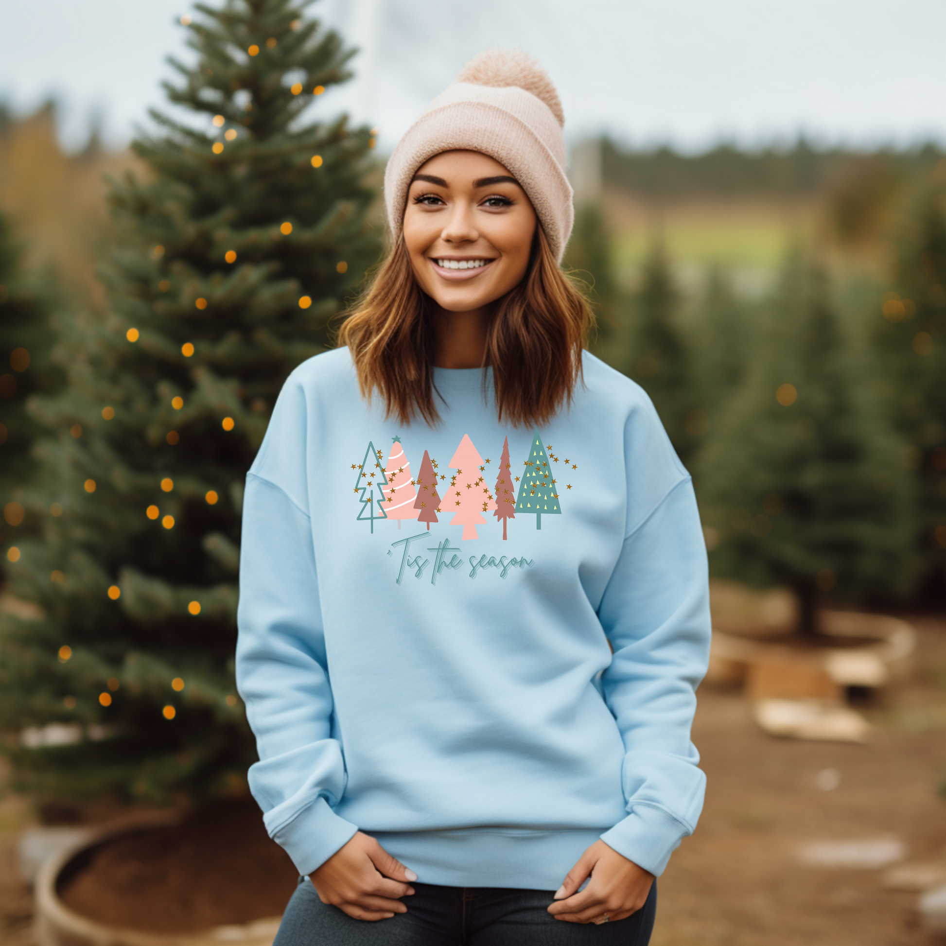 A cozy woman wearing a Light Blue Christmas Tree Sweatshirt by Printify posing in front of Christmas trees.