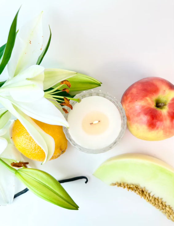 A Good Vibes Woodwick Candle by Aroma Avenu, surrounded by a watermelon slice, lemon slice, and lily flower on a white background, radiating positive energy for good vibes.