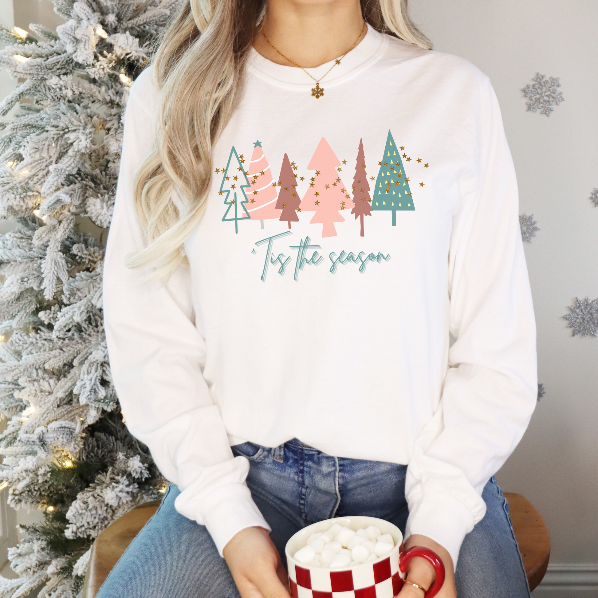 A woman, dressed in her cozy and stylish winter wardrobe, is sitting in front of a Christmas tree with a cup of hot cocoa while wearing the Women's 'Tis the Season White Christmas Tree Shirt by Printify | Comfort Colors Holiday Tee.