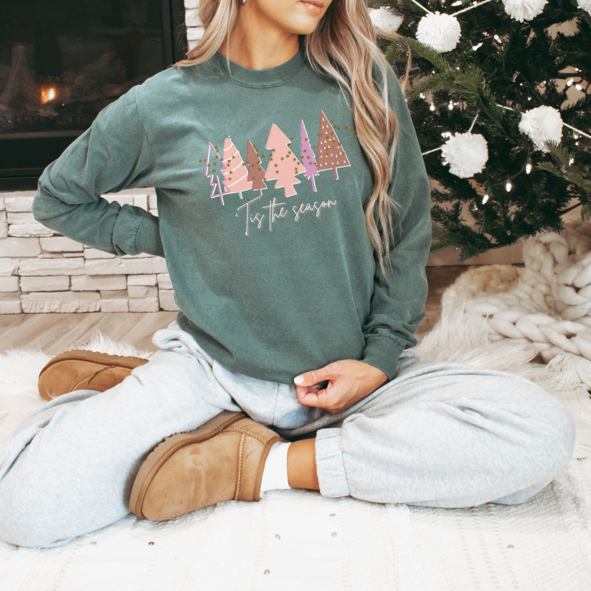 A woman sitting in front of a Christmas tree wearing a Women's 'Tis the Season Light Green & Pink Christmas Tree Shirt | Comfort Colors Holiday Tee by Printify, adding a touch of comfort and style to her winter wardrobe.