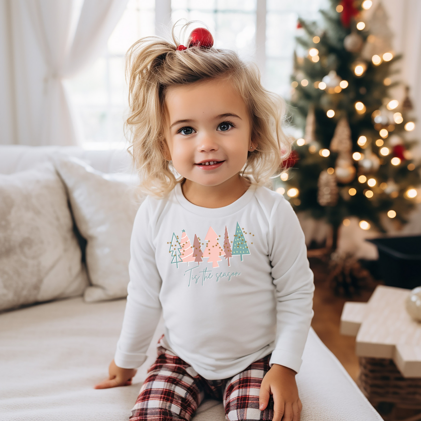 A little girl wearing a Kids Christmas Tree White Long Sleeve Tee - Toddler Crew Neck T-Shirt by Printify sitting on a couch in front of a Christmas tree at an Etsy store.