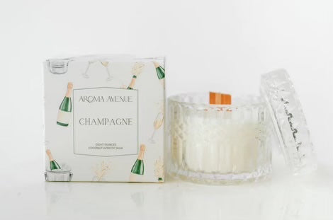The enticing aroma of Aroma Avenu Champagne Woodwick Candle fills the room, complemented by the presence of a bottle of champagne.