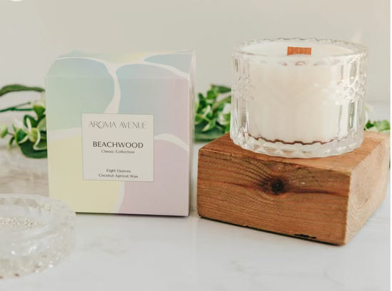 The Beachwood Woodwick Candle from Aroma Avenue, with its tropical coconut twist, is sitting on a wooden block next to a plant.