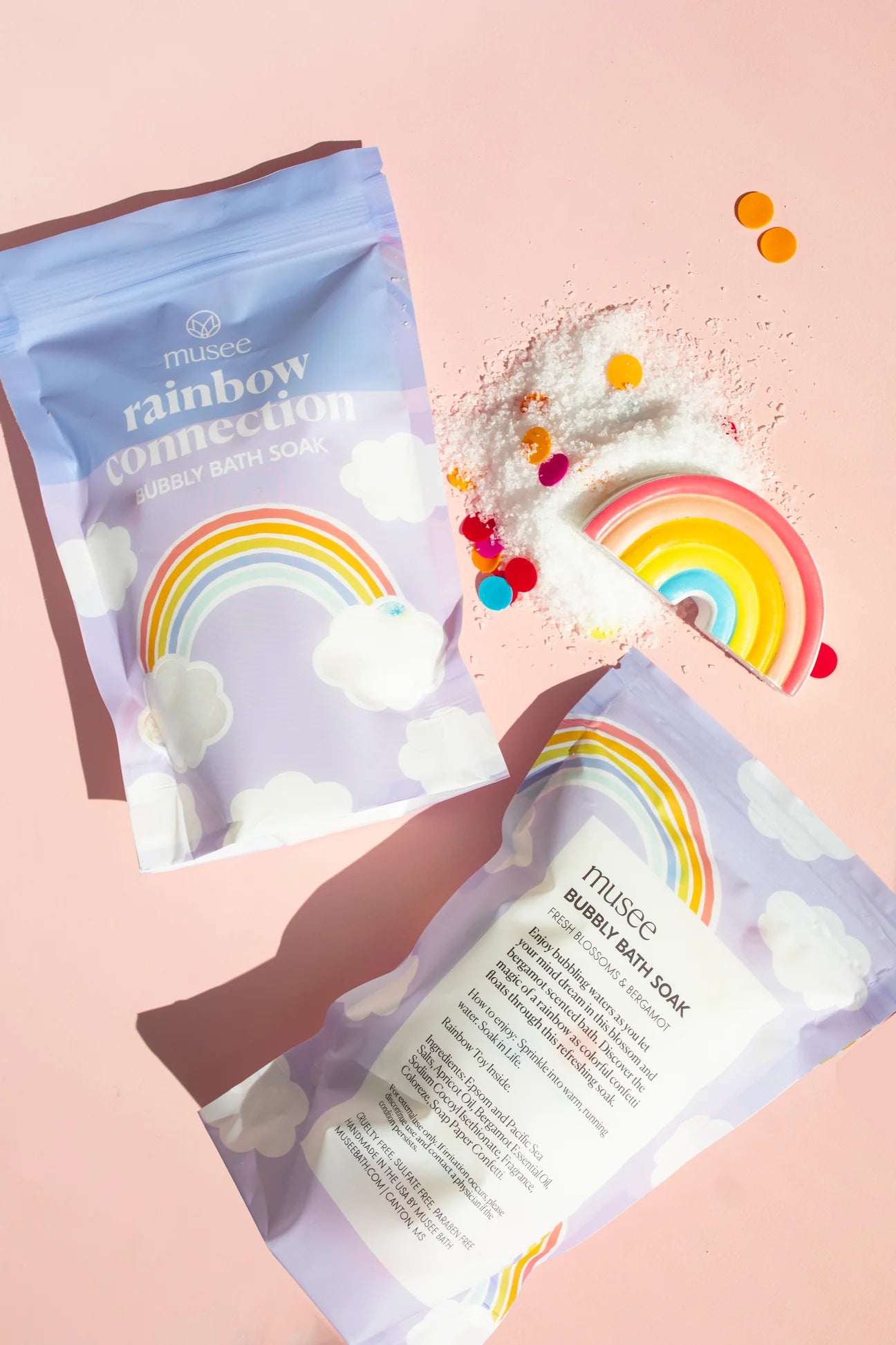 A bag of Rainbow Connection Bubbly Bath Soak and a bag of sprinkles creating a magical oasis on a pink background. (Brand: Musee)