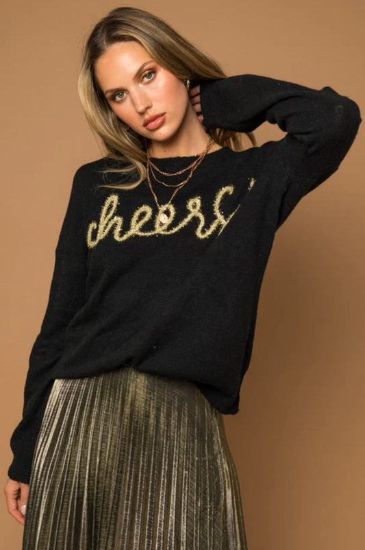 A woman wearing a "Cheers" pullover sweater by Chickie Collective, and a gold plea skirt.