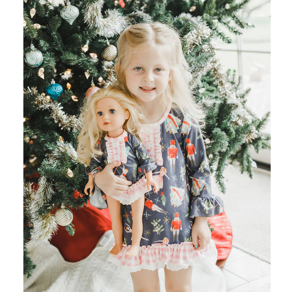 A little girl holding a Nutcracker Set with Pink Gingham doll in front of a christmas tree by Chickie Collective.
