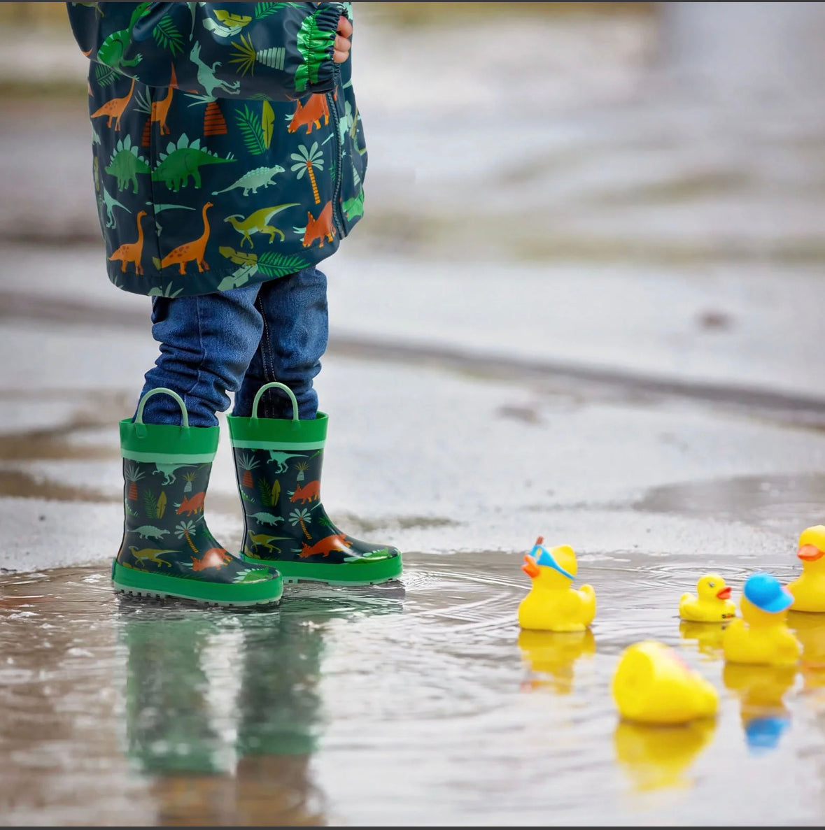 A child is standing in a puddle with Stephen Joseph's Rain Boots - Green Dinosaur.