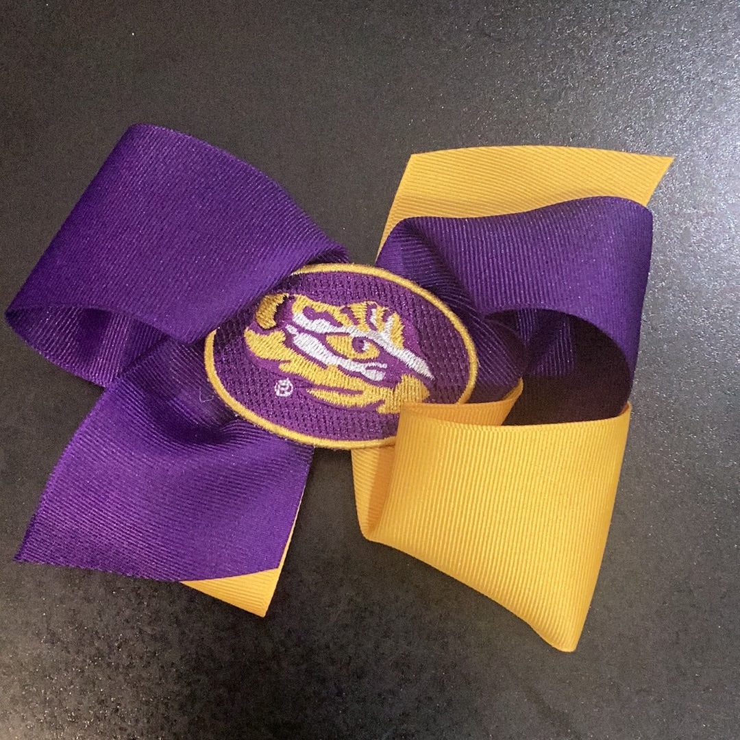 A large purple and yellow LSU bow with an embroidered eye of the tiger in the middle, featuring a no-slip clip for easy securing by Wee Ones.