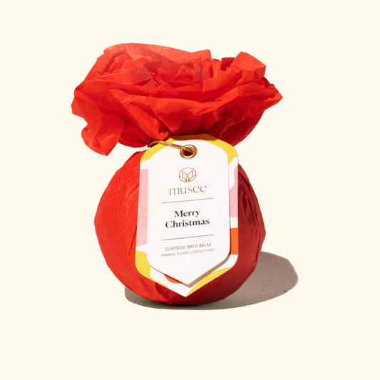 A red holiday gift bag with a Musee Merry Christmas Surprise Bath Balm tag on it.