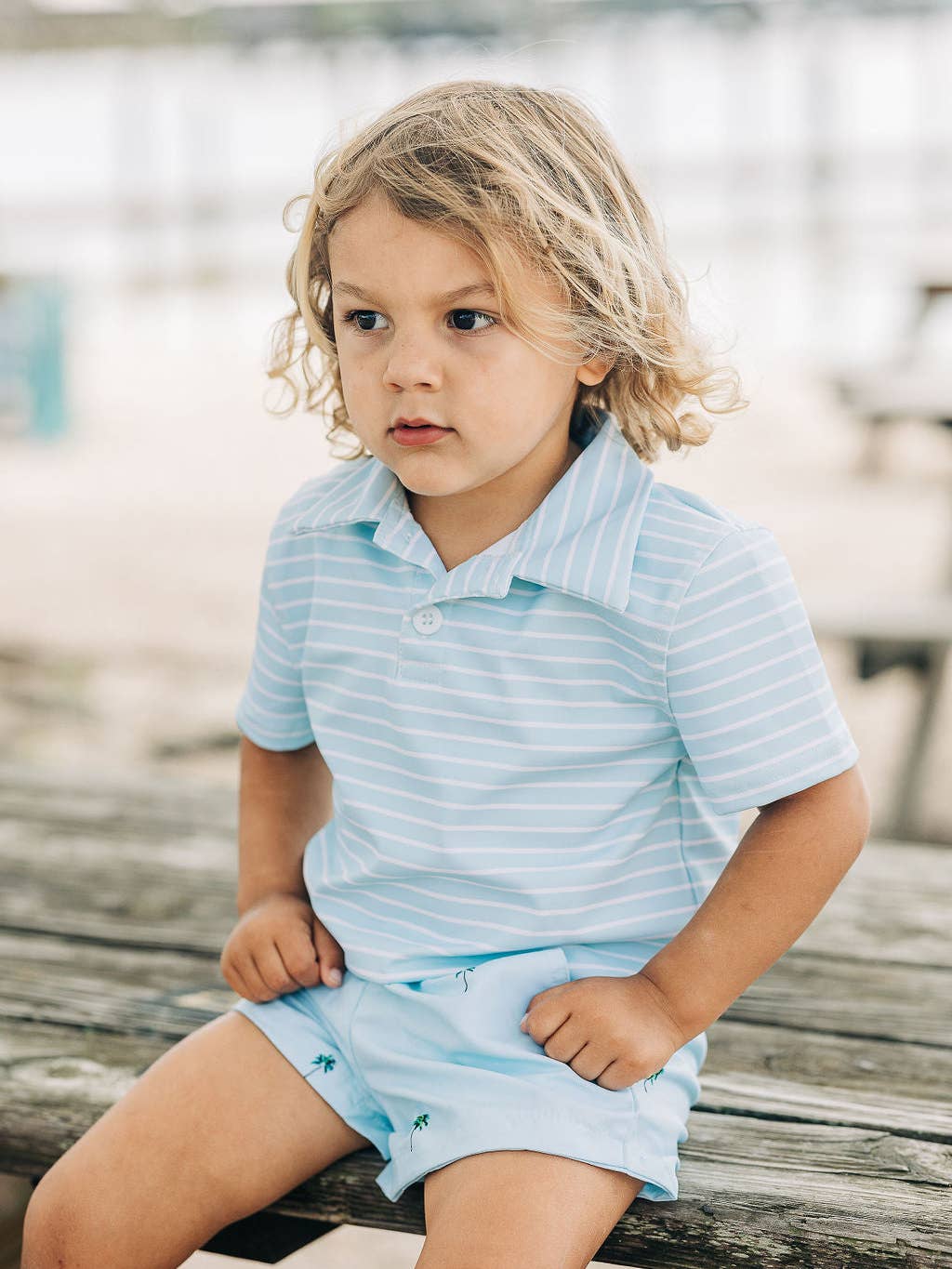 a little boy wearing a Sugar Bee Clothing Blue Stripe Polo Shirt and sitting on a wooden bench.