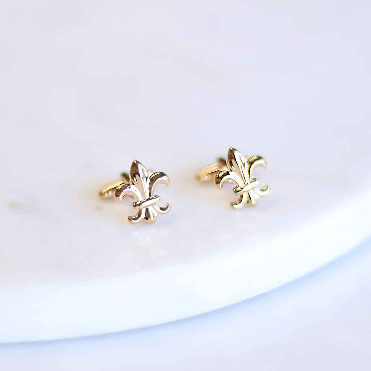 A pair of The Royal Standard Fleur De Lis Cufflinks in gold, measuring .5", on a white plate.