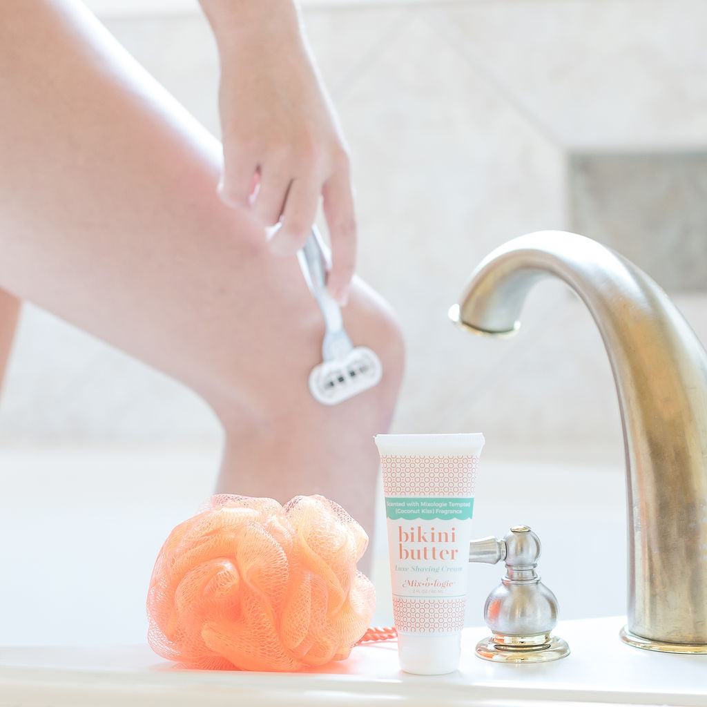 A woman is using Mixologie's Bikini Butter to shave her legs in a bathtub.