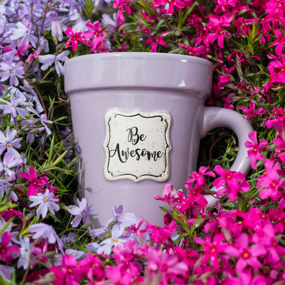 A Nicole Brayden Lilac Flower Pot Mug with a sticker that says Be Awesome surrounded by purple flowers.
