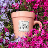 A Peach Flower Pot Mug - You Are Loved by Nicole Brayden sitting in a field of purple flowers.