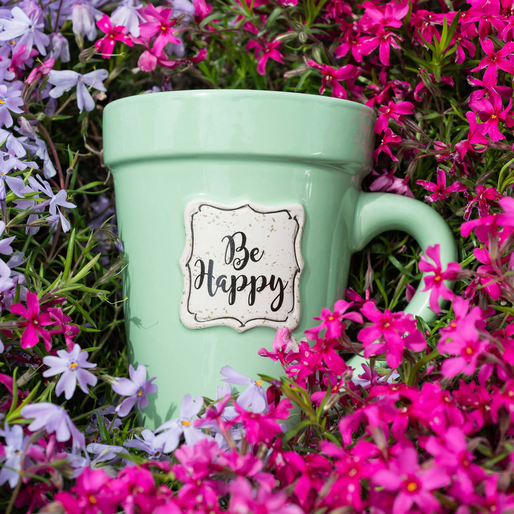 A Nicole Brayden Green Flower Pot Mug - Be Happy with a be happy sticker on it surrounded by purple flowers.