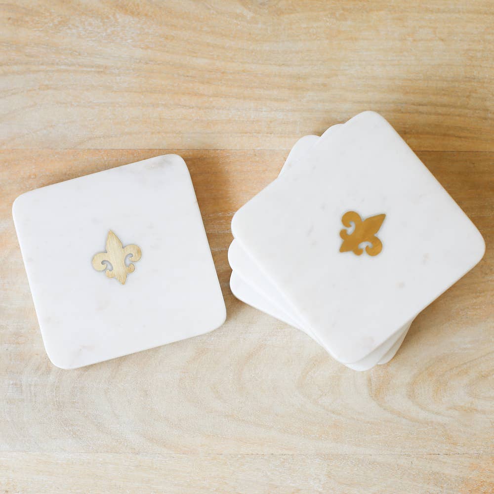 A couple of Fleur de Lis Marble Coasters in White/Brass 4x4 from The Royal Standard sitting on top of a wooden table.