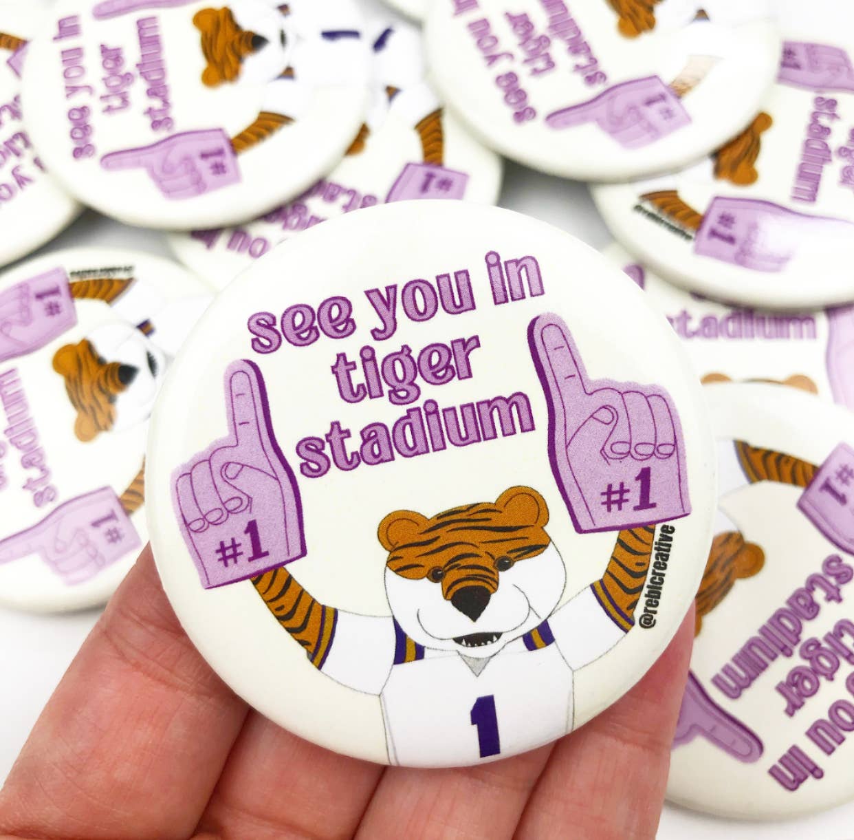 With enthusiastic team spirit, a person is proudly holding a "GAME DAY BUTTON - See you in Tiger Stadium" that bears the brand name of REBL Creative.