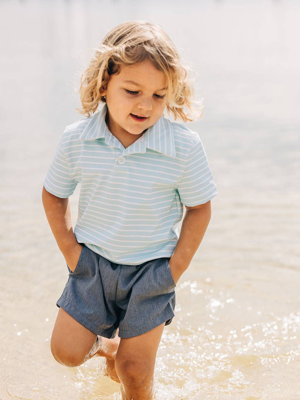 a little boy wearing a Sugar Bee Clothing Blue Stripe Polo Shirt standing in some water.