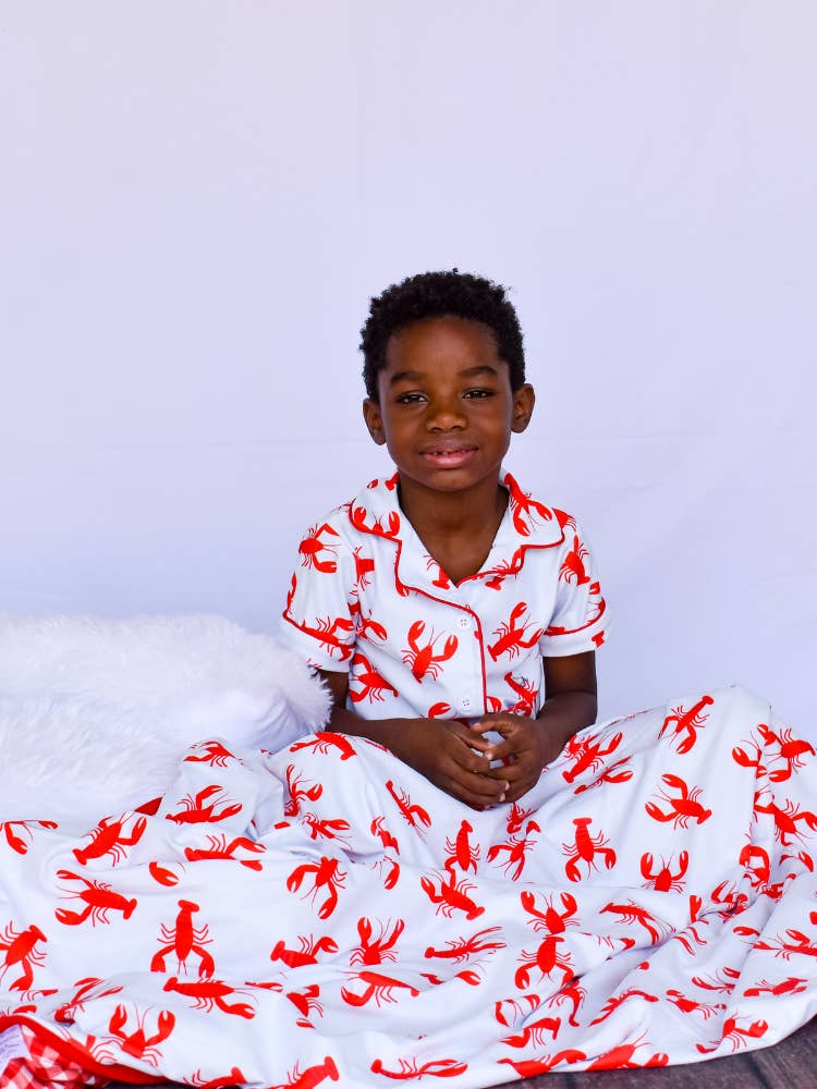A young boy in Sugar Bee Clothing's Crawfish Button Down Pajamas sitting on a bed.